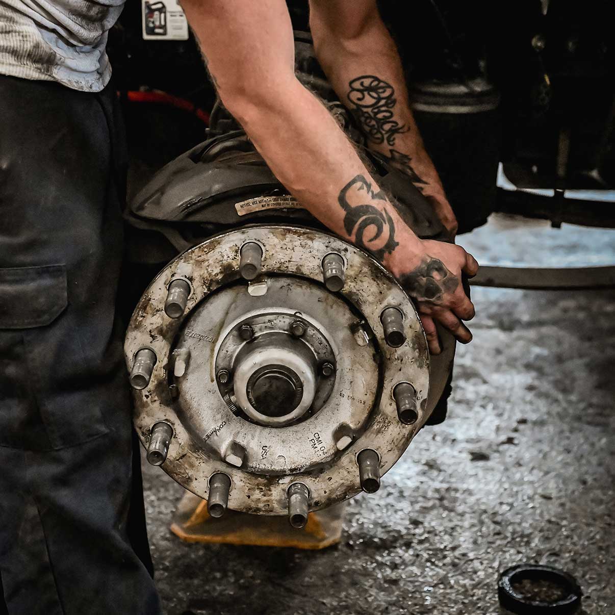 A person with tattooed arms is working on a large vehicle wheel hub in a workshop, holding and inspecting the metal part. The workshop floor is dirty and tools are visible in the background.