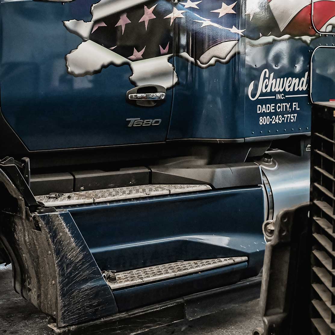 Side view of a dark blue truck with a ripped American flag graphic and “Schwend Inc. DADE CITY, FL” with a phone number printed on the door. The truck's steps and part of the cab are visible.