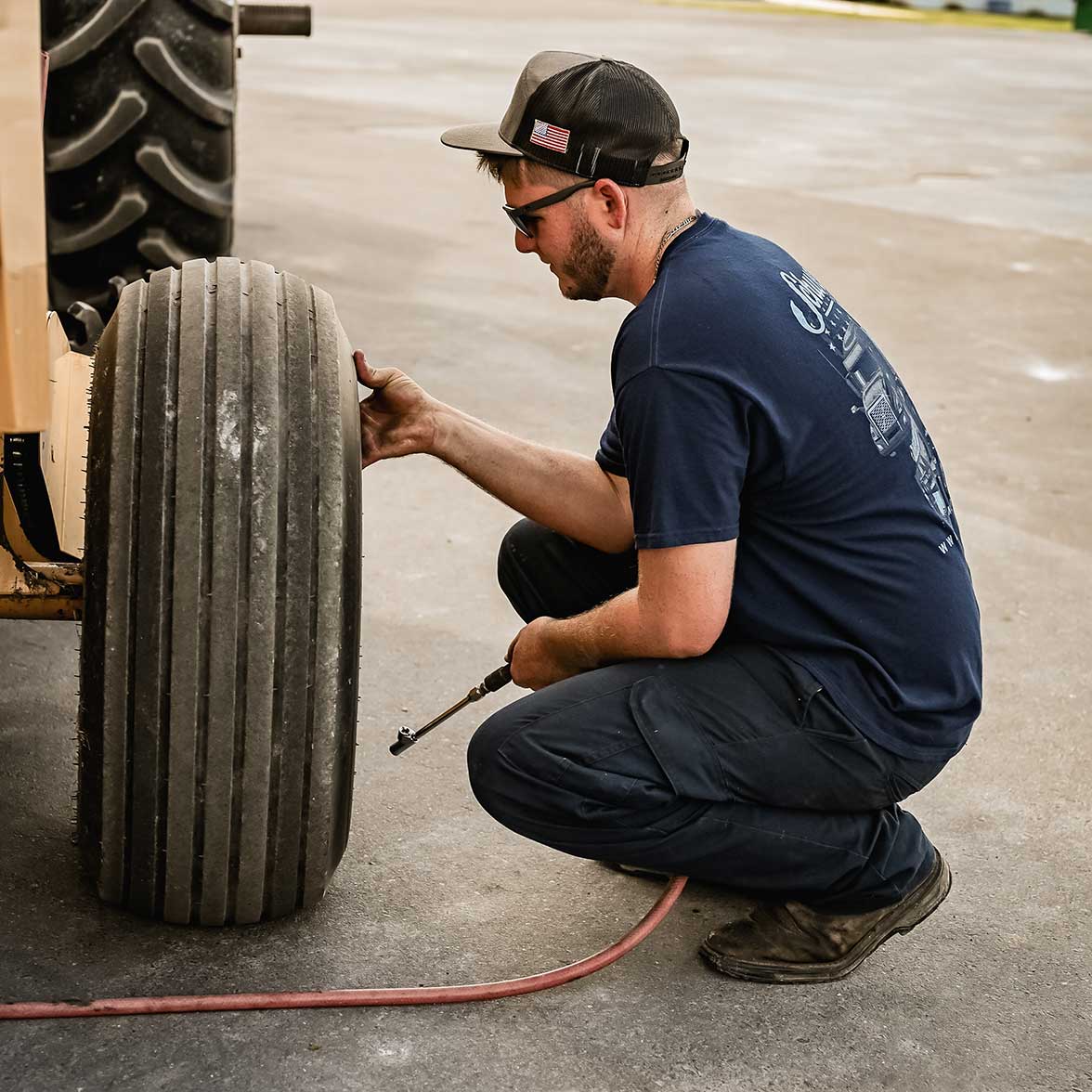 A man wearing a navy t-shirt, cap, and sunglasses kneels on a concrete surface, checking the air pressure of a large vehicle tire with a pressure gauge and hose.