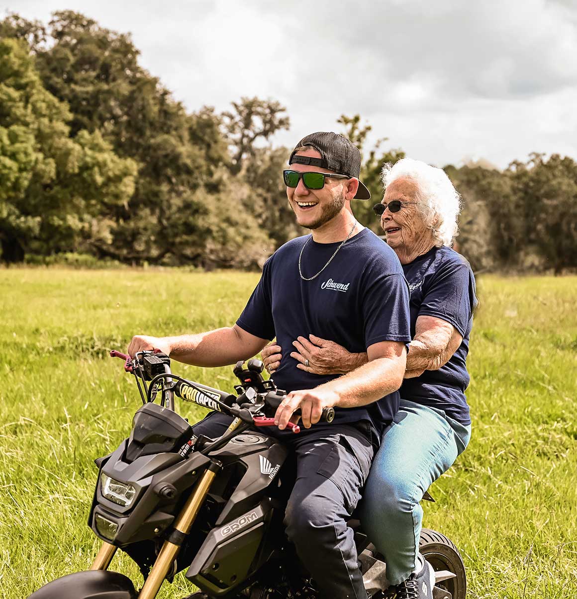 A young man and an elderly woman ride together on a motorcycle through a grassy field, both smiling and enjoying the moment on a sunny day.